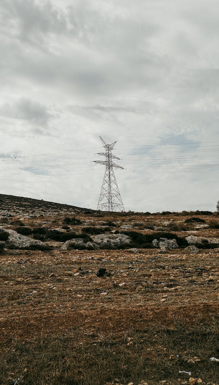 A high-voltage transmission tower set against a rocky rural landscape and cloudy sky.